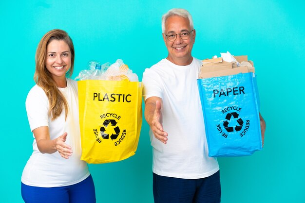 Pareja de mediana edad sosteniendo bolsas de reciclaje llenas de papel y plástico aislado sobre fondo blanco un apretón de manos para cerrar un buen trato