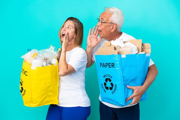 Pareja de mediana edad sosteniendo bolsas de reciclaje llenas de papel y plástico aislado sobre fondo blanco gritando con la boca abierta hacia el lateral