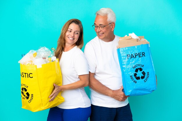 Pareja de mediana edad sosteniendo bolsas de reciclaje llenas de papel y plástico aislado sobre fondo blanco mirando por encima del hombro con una sonrisa