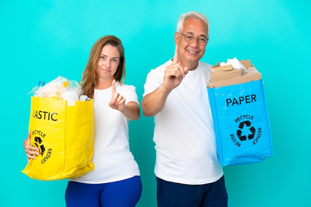 Pareja de mediana edad sosteniendo bolsas de reciclaje llenas de papel y plástico aislado sobre fondo blanco mostrando y levantando un dedo