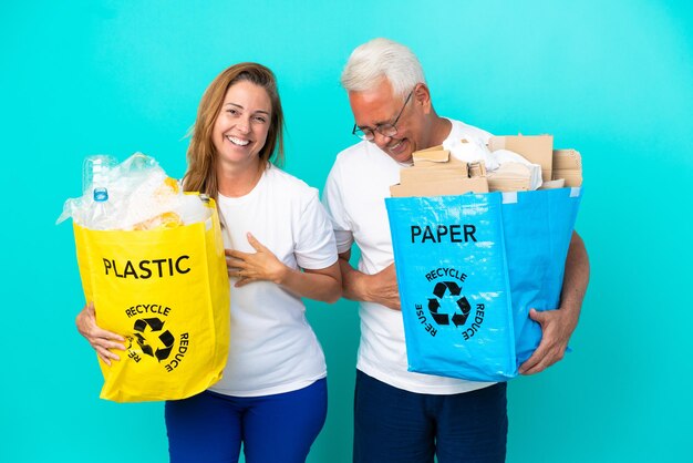 Pareja de mediana edad sosteniendo bolsas de reciclaje llenas de papel y plástico aislado sobre fondo blanco sonriendo mucho mientras pone las manos en el pecho