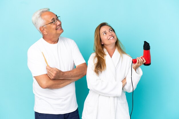 Pareja de mediana edad sosteniendo secador y cepillo de dientes aislado sobre fondo azul mirando hacia arriba mientras sonríe