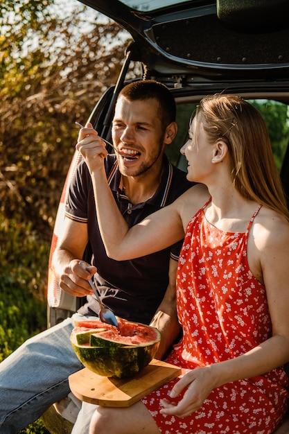 Pareja milenaria sentada en el baúl abierto y comiendo sandía Feliz pareja joven teniendo un descanso en el campo