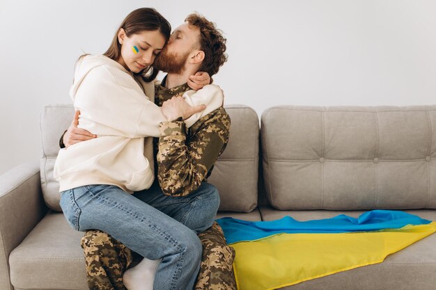 Una pareja de militares ucranianos uniformados besa a su novia en el sofá de casa con un fondo de bandera amarilla y azul