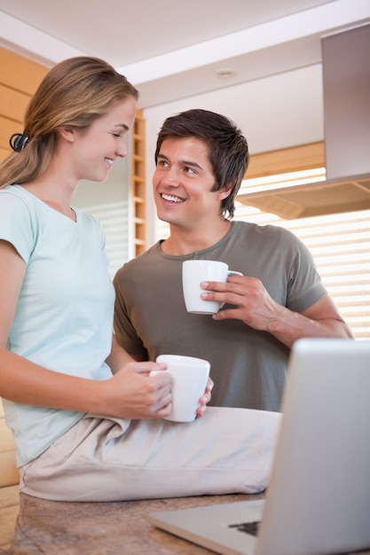 Pareja mirando cada uno con tazas de café mientras usa la computadora portátil