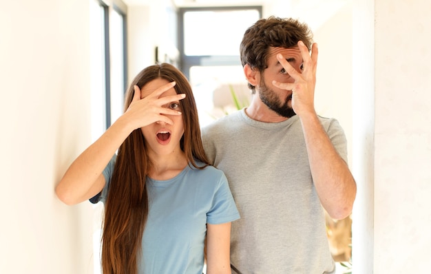 Pareja mirando conmocionado, asustado o aterrorizado, cubriendo la cara con la mano y mirando entre los dedos