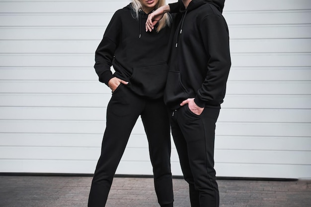 Una pareja de moda posa al aire libre con sudaderas negras con capucha para un diseño de maqueta una plantilla de moda para impresión y marca un estilo de ropa de calle casual y elegante sin cara y sin logotipo visible