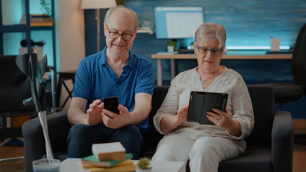 Pareja moderna usando smartphone y tableta digital en casa. Personas mayores con discapacidad física mirando teléfonos móviles y aparatos para divertirse con la tecnología en la sala de estar. familia mayor