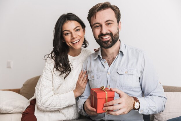 Pareja morena hombre y mujer sosteniendo caja de regalo de cumpleaños mientras está sentado en el sofá en una habitación luminosa en casa