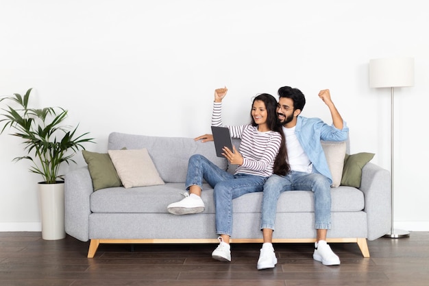 Pareja multicultural emocional sentada en un sofá usando una tableta digital