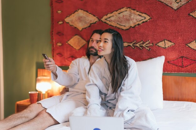 Pareja multiétnica en albornoces en la cama viendo la televisión y usando una computadora portátil