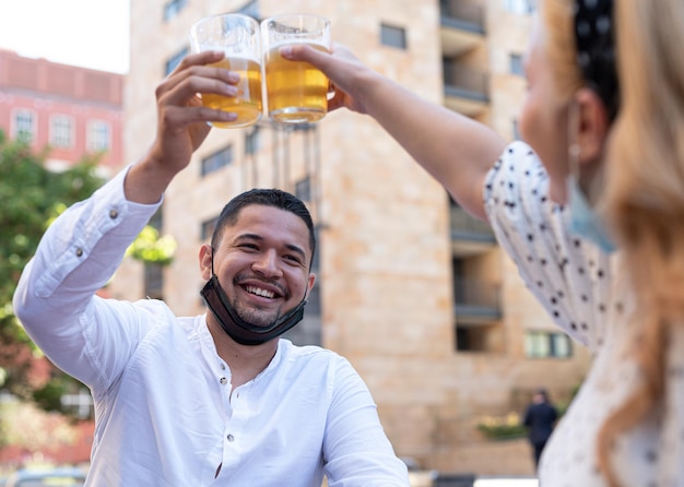 Foto pareja multiétnica brindando y celebrando, retrato de la sonrisa feliz del hombre con la máscara quirúrgica debajo de la barbilla, la gente comienza a socializar nuevamente después del encierro