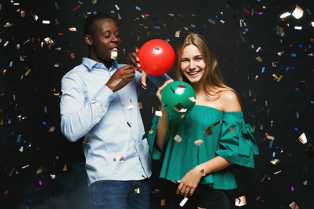 Pareja multiétnica divirtiéndose. Hombre y mujer joven con globos en fondo negro, bañados con confeti. Concepto de celebración de fiesta de cumpleaños o año nuevo