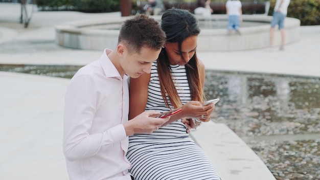 Pareja multiétnica sentados juntos al aire libre y mirando en los teléfonos inteligentes