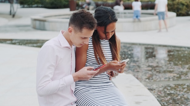 Pareja multiétnica sentados juntos al aire libre y usando teléfonos inteligentes