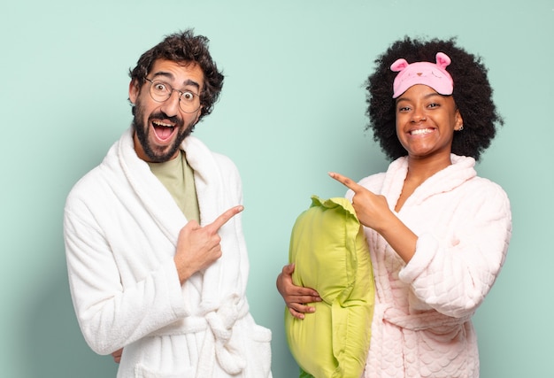 Foto pareja multirracial de amigos que parecen emocionados y sorprendidos apuntando hacia un lado y hacia arriba para copiar el espacio. pijamas y concepto de hogar.