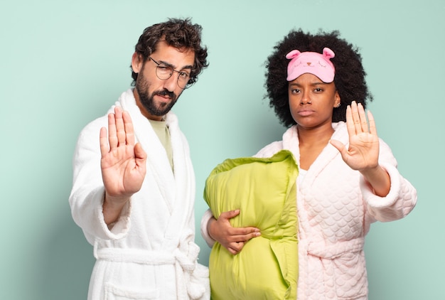 Pareja multirracial de amigos que parecen serios, severos, disgustados y enojados mostrando la palma abierta haciendo un gesto de parada. pijamas y concepto de hogar.