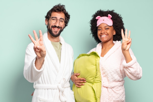 Pareja multirracial de amigos sonriendo y mirando amistosamente, mostrando el número tres o tercero con la mano hacia adelante, contando hacia atrás. pijamas y concepto de hogar.