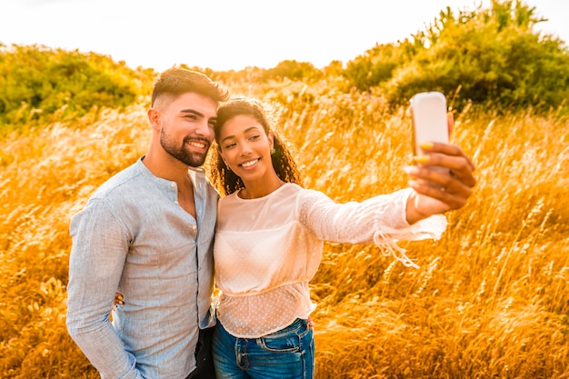 Pareja multirracial enamorada divirtiéndose con smartphone tomando selfie en la naturaleza. Chica hispana se fotografía con su novio al atardecer para compartir vacaciones en las redes sociales. Gente joven hermosa