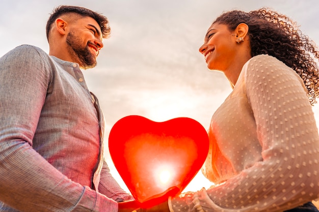 Pareja multirracial de enamorados celebrando el Día de San Valentín mirando a los ojos el uno al otro cogidos de la mano con un globo rojo en forma de corazón atravesado por los rayos del sol poniente. Vista desde la parte inferior de los jóvenes amantes coqueteando