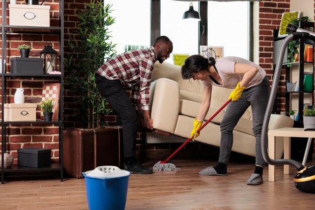 Pareja multirracial haciendo limpieza de primavera en casa, esposo afroamericano levanta un pesado sofá de la sala para que la esposa limpie debajo. Pareja cuidando juntos las tareas del hogar.