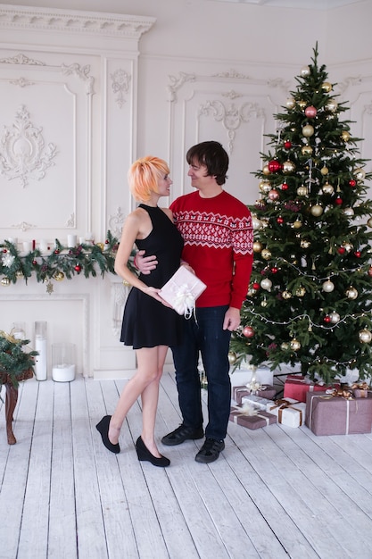 Pareja en Navidad con caja de regalos en su sala de estar, frente al árbol de Navidad