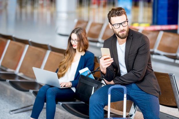 Pareja de negocios elegante sentado con laptop, teléfono y maleta en la sala de espera en el aeropuerto. Concepto de viajes de negocios