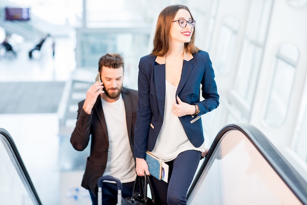 Pareja de negocios hablando con teléfonos mientras se sube a las escaleras mecánicas con equipaje en el aeropuerto