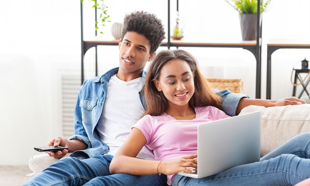Una pareja negra se relaja en un sofá en su casa la chica está usando una computadora portátil mientras el chico