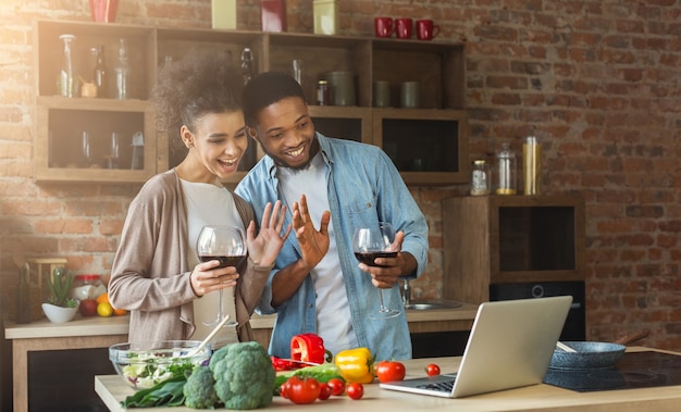 Foto pareja negra usando laptop y saludando mientras bebe vino en la cocina tipo loft