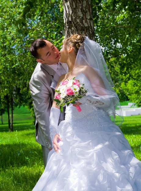 La pareja de novios se besa cerca del tronco de un árbol.