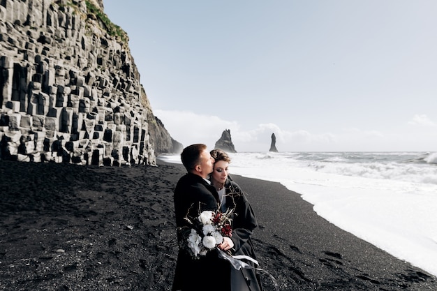 Una pareja de novios camina por la playa de arena negra de vik cerca de la roca basáltica