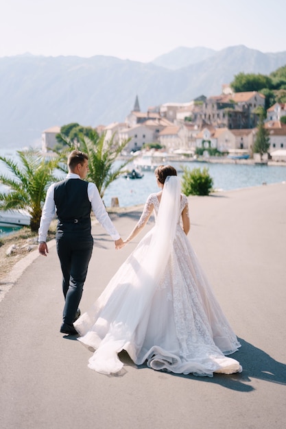 Pareja de novios camina por el terraplén cerca del mar, con el telón de fondo de palmeras, montañas y la ciudad vieja