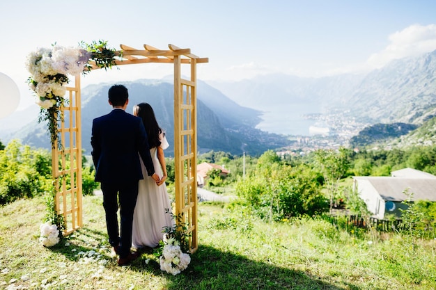 Pareja de novios de luna de miel viaje vista posterior Arco de boda montañas y fondo marino en Montenegro Estilo de boda de bellas artes