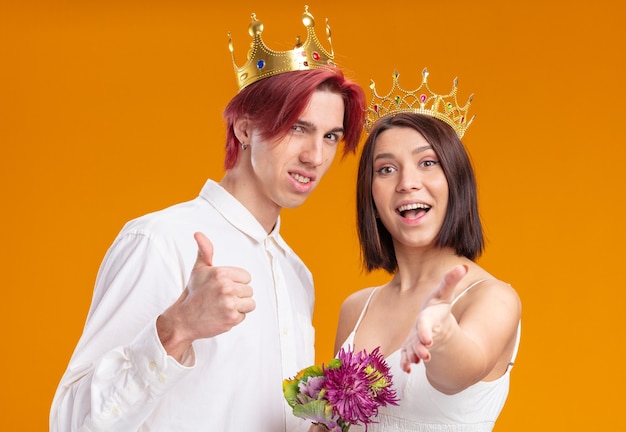 Pareja de novios con ramo de flores en vestido de novia con coronas de oro sonriendo alegremente posando juntos mostrando thums de pie sobre la pared naranja
