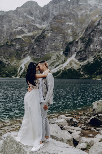 Pareja de novios románticos en el amor de pie en la orilla pedregosa del lago Sea Eye en Polonia. Vista panorámica a la montaña. La novia y el novio. Morskie Oko. Montes Tatra.