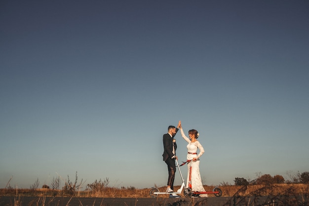 Pareja de novios sonriente montando en scooters a lo largo de la carretera fuera de la ciudad al atardecer.