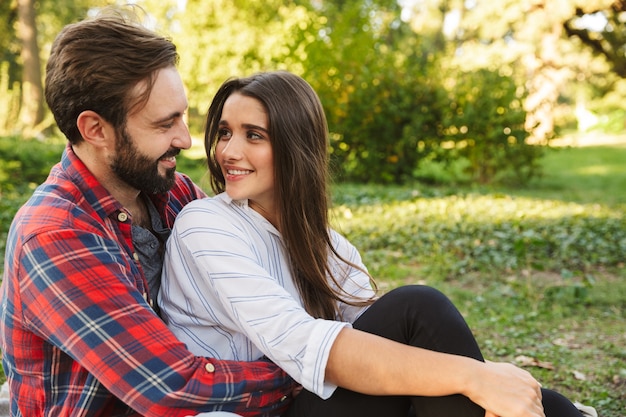 Pareja optimista hombre y mujer vestidos con ropa casual abrazándose y mirándose mientras descansa en el parque verde