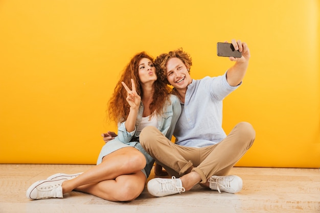 Pareja optimista joven y mujer sentados en el piso juntos y mostrando el signo de la paz mientras toman selfie en el teléfono inteligente, aislado sobre fondo amarillo