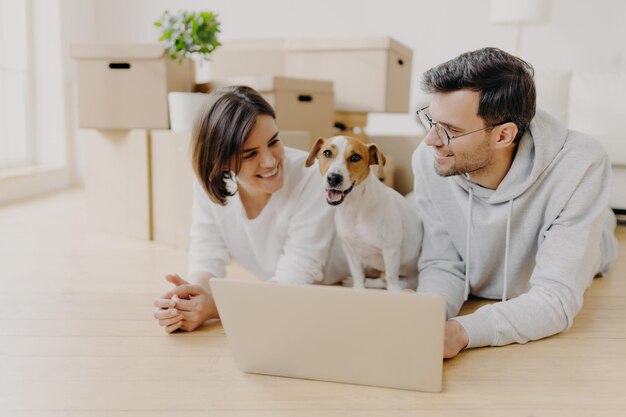 Una pareja con un perro usando una computadora portátil mientras yacen en el suelo de una casa nueva