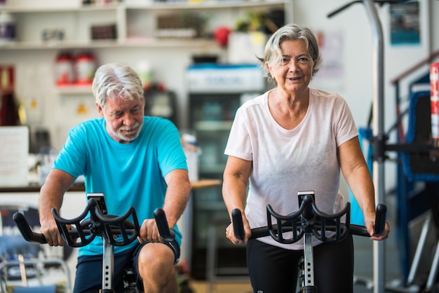 Pareja de personas maduras o personas mayores activas que hacen ejercicio en el gimnasio montando la bicicleta en el interior para estar saludables: dos jubilados felices con un estilo de vida saludable