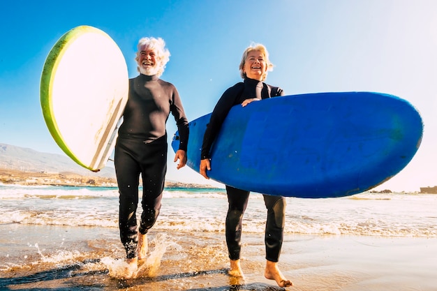 Foto pareja de personas mayores en la playa con trajes de neopreno negros sosteniendo una mesa de surf lista para ir a surfear en la playa: personas maduras y jubiladas activas que realizan actividades felices juntas en sus vacaciones o tiempo libre