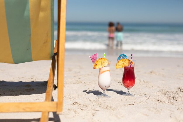 Una pareja de raza mixta disfruta del tiempo libre en la playa en un día soleado junto con cócteles en la arena en primer plano.