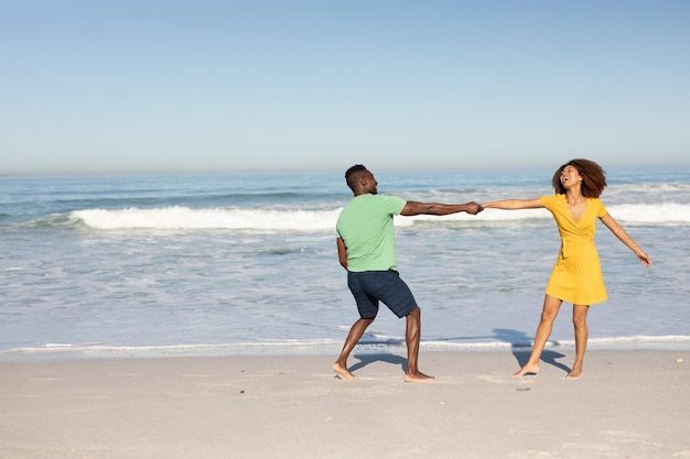 Una pareja de raza mixta disfrutando juntos del tiempo libre en la playa en un día soleado, bailando y riendo con el sol brillando en sus rostros. Relajantes vacaciones de verano.