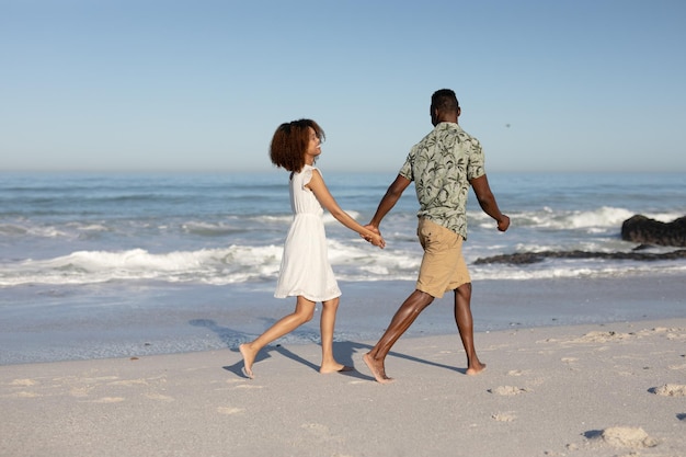 Una pareja de raza mixta disfrutando juntos del tiempo libre en la playa en un día soleado, caminando y abrazándose con el sol brillando en sus rostros.