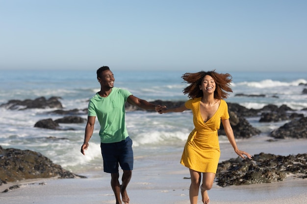 Una pareja de raza mixta disfrutando juntos del tiempo libre en la playa en un día soleado, tomándose de la mano, divirtiéndose y riéndose con el sol brillando en sus caras. Relajantes vacaciones de verano.