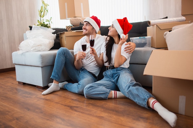 La pareja de recién casados celebra la Navidad o el Año Nuevo en su nuevo apartamento. Joven feliz y mujer bebiendo vino, celebrando mudarse a casa nueva y sentados entre cajas.