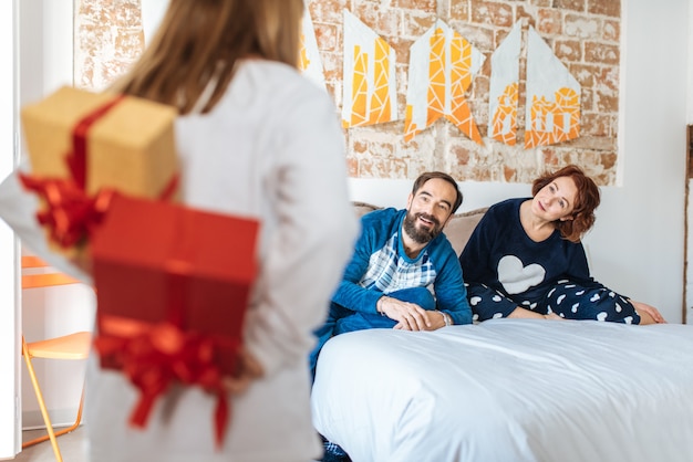 Pareja relajada en casa en la cama con pequeña hija haciendo una sorpresa con regalos