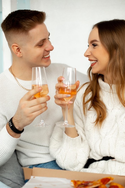 Una pareja riéndose compuesta por un hombre rubio con chaqueta blanca y una mujer romántica vestida con un cárdigan blanco con cinturón negro tintineando copas de champán mientras celebran el día de san valentín