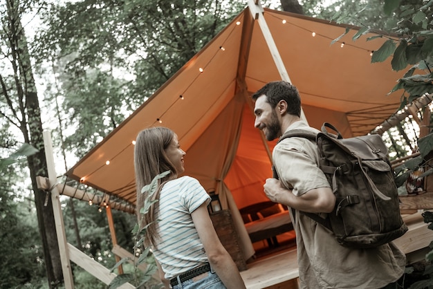 Pareja romántica acampando al aire libre y de pie en la tienda glamping. Feliz el hombre y la mujer en unas románticas vacaciones de camping. Hombre con mochila con su novia millennials llegó al sitio de glamping.